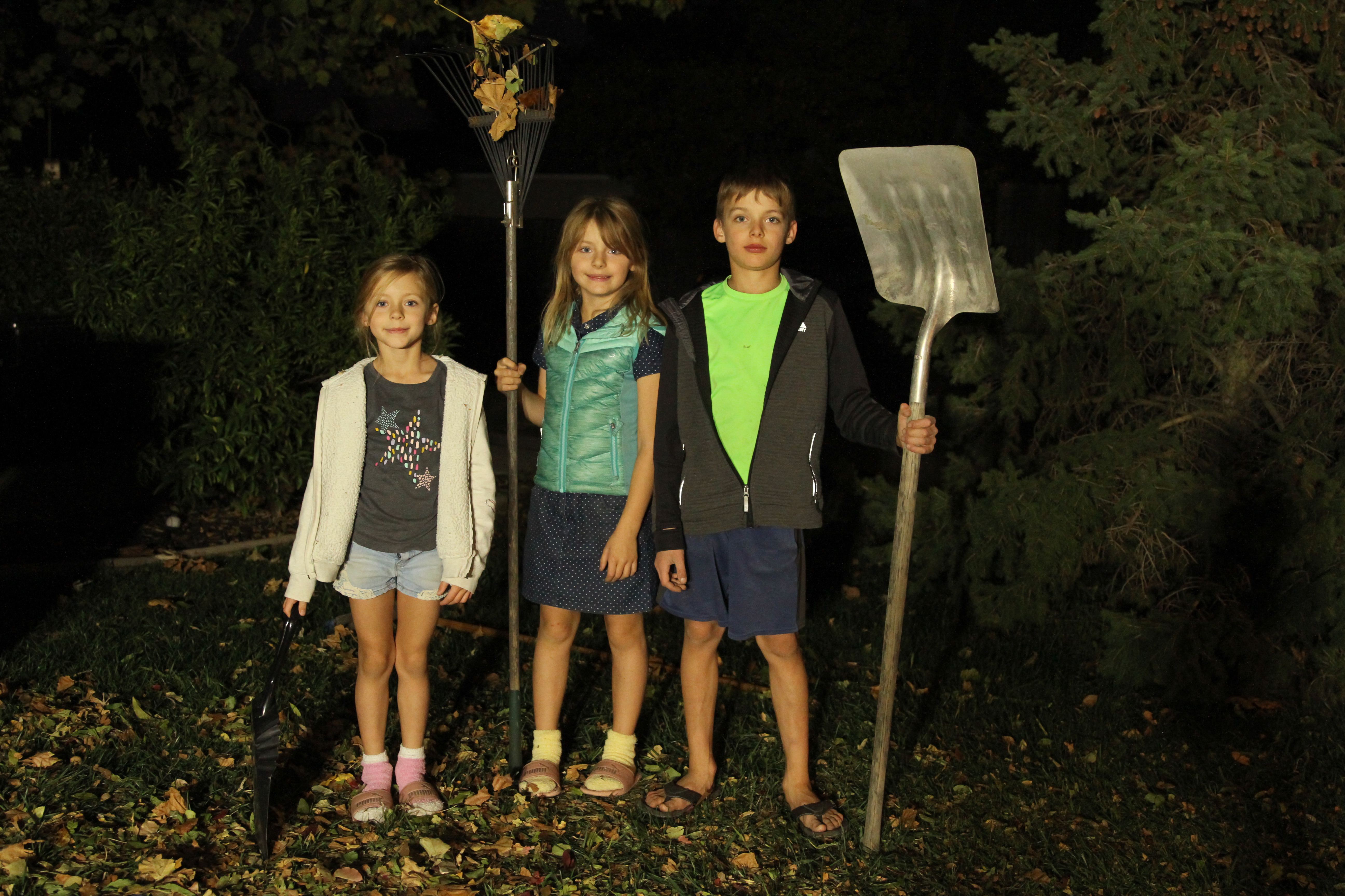 3 kids raking leaves in the fall posing with rakes and shovels